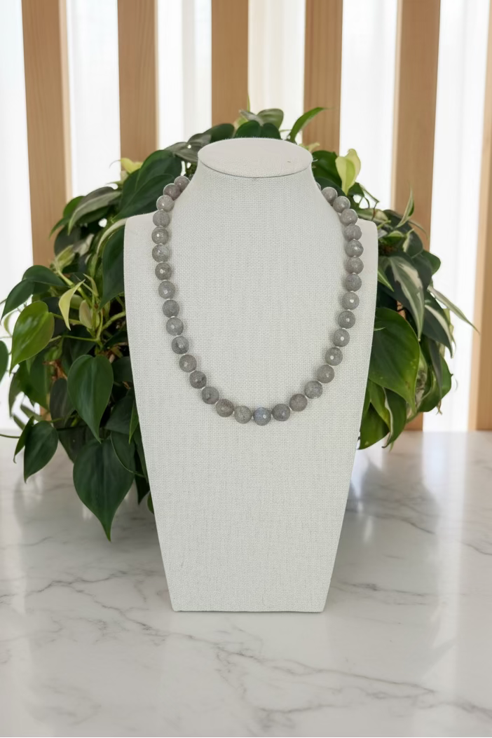 A gray beaded necklace displayed on a white fabric jewelry bust, set on a marble table with green plants in the background.