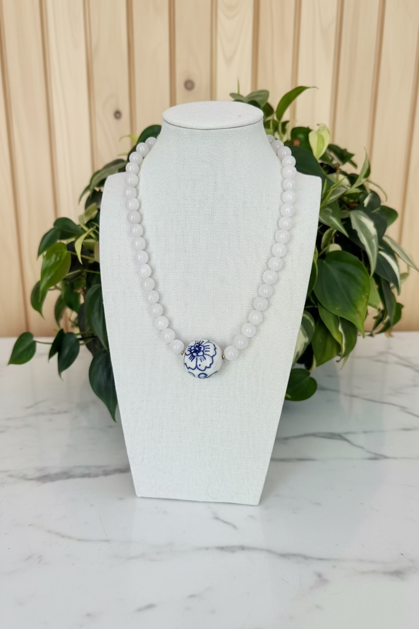 A white beaded necklace with a central blue and white floral bead, displayed on a white fabric jewelry bust with green plants in the background.