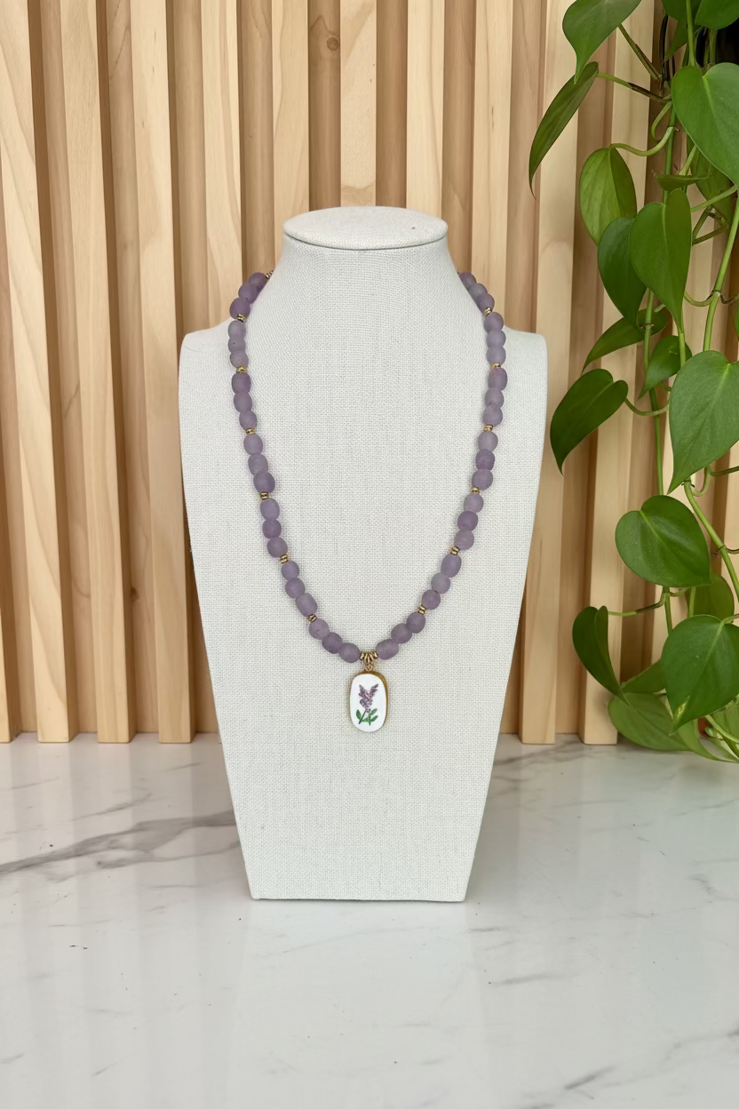 A purple african beaded necklace with a floral rectangular pendant displayed on a white fabric mannequin bust on a wooden table with green plants in the background.
