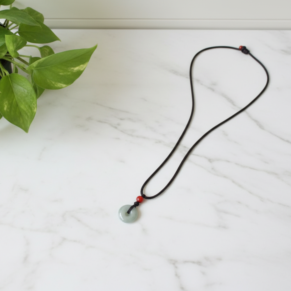 Necklace with a green jade pendant and a brown cotton cord on a white marble counter, with a plant in the background