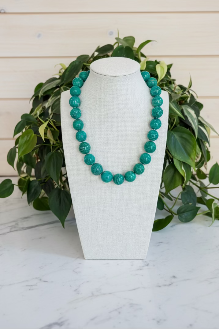 A beaded necklace made of large, polished teal-green round beads displayed on a white fabric mannequin bust, set on a marble table with green plants in the background.