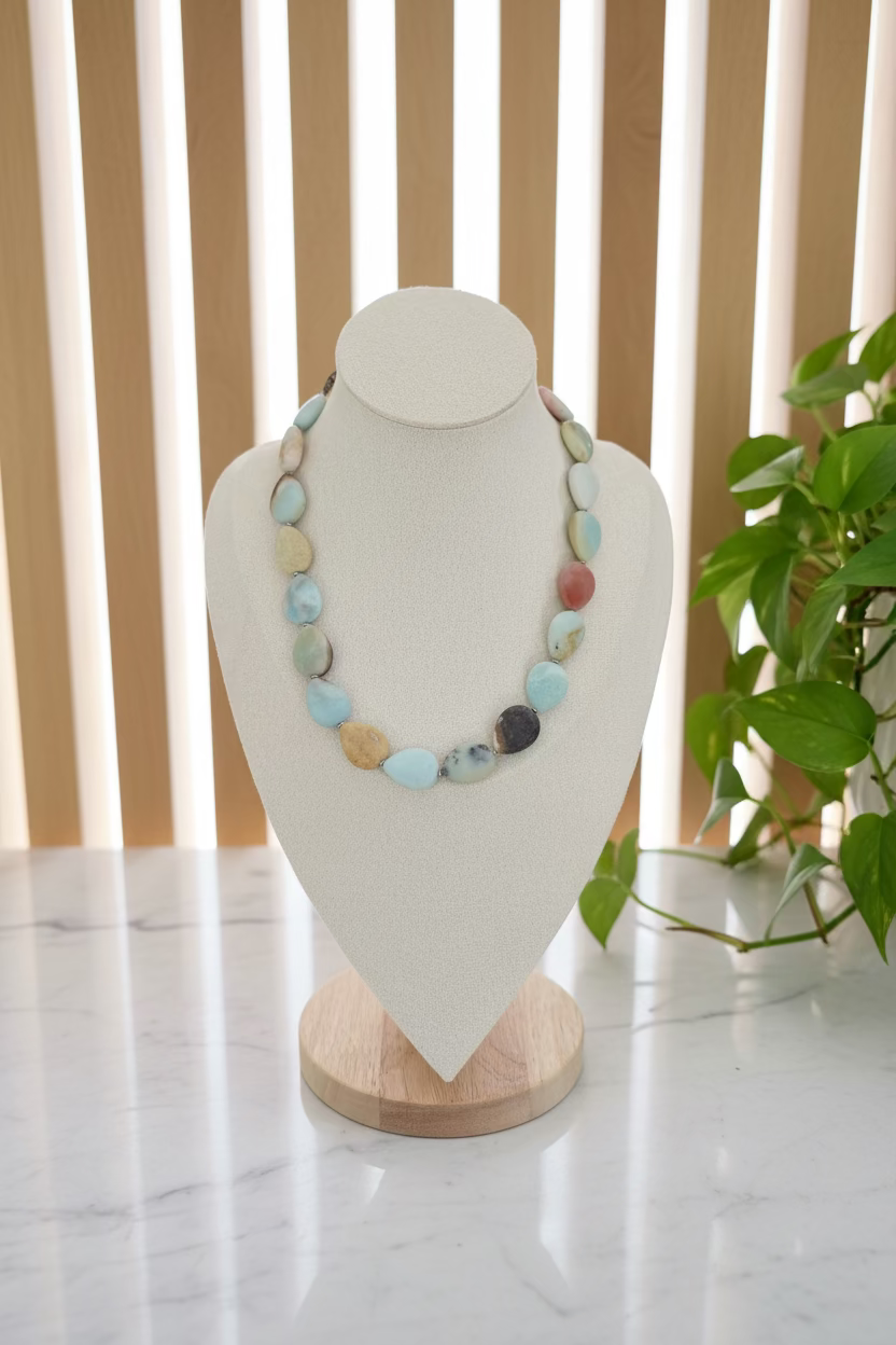 A multi-colored stone bead necklace displayed on a cream fabric jewelry bust. The necklace features smooth, polished oval and teardrop-shaped beads in various colors including light blue, beige, green, brown, and reddish-brown, strung on a light wood stand.