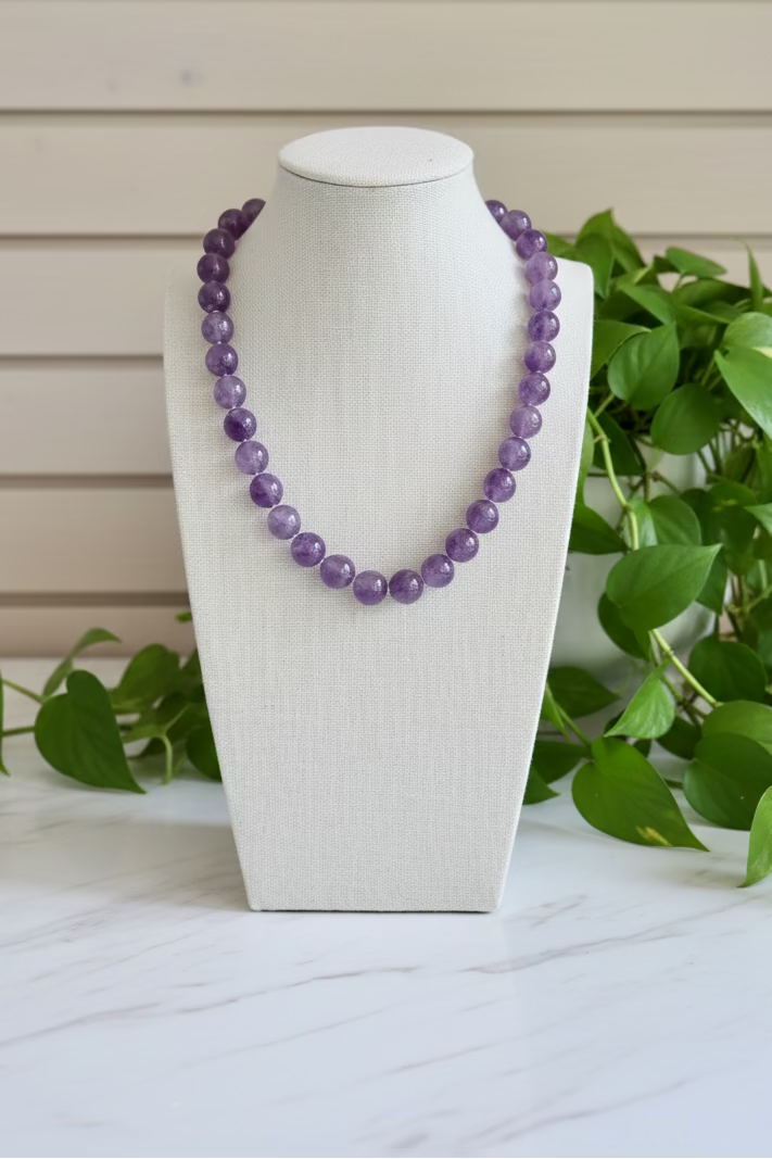 A purple beaded necklace displayed on a white fabric-covered jewelry bust, set on a marble table with green plants 