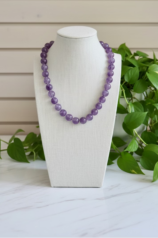 A purple beaded necklace displayed on a white fabric-covered jewelry bust, set on a marble table with green plants 