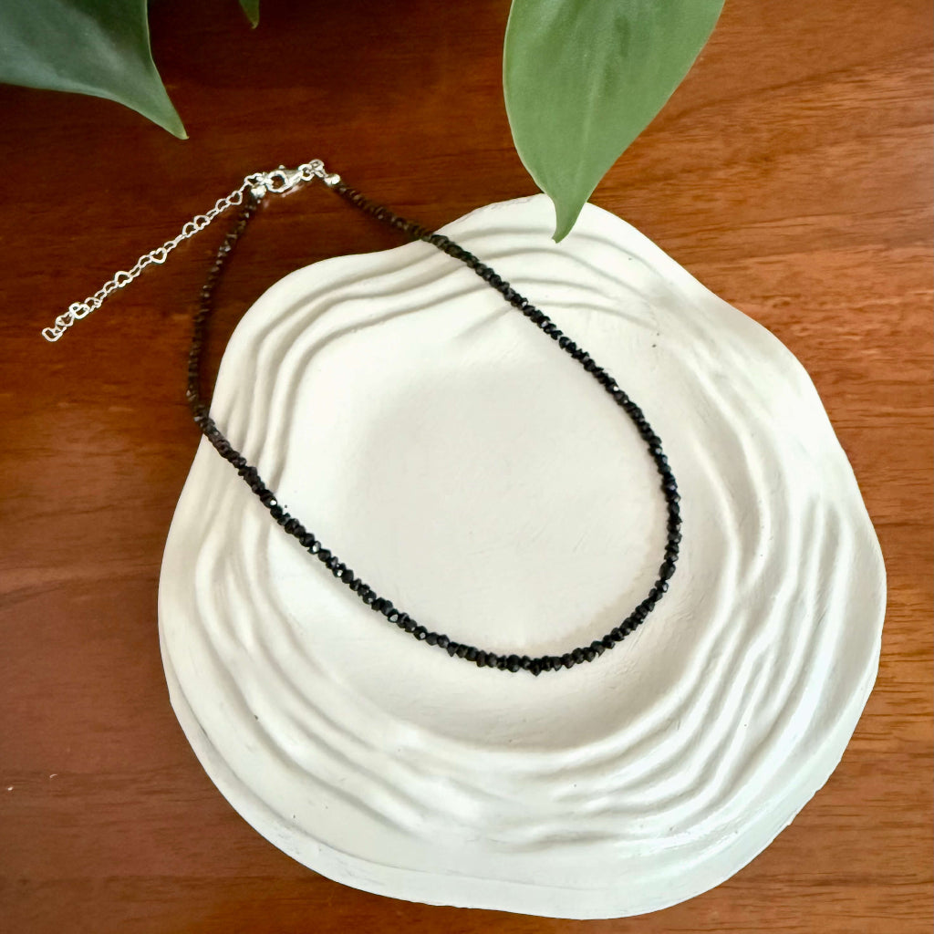 Black spinel beaded gemstone necklace on a white ceramic dish with green leaves in the background