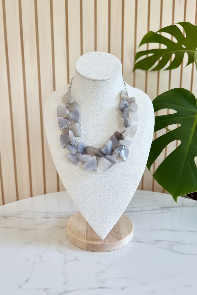 A chunky statement necklace made of irregular light gray and lavender agate-like stone chips, displayed on a white fabric jewelry bust against a background of green houseplants.
