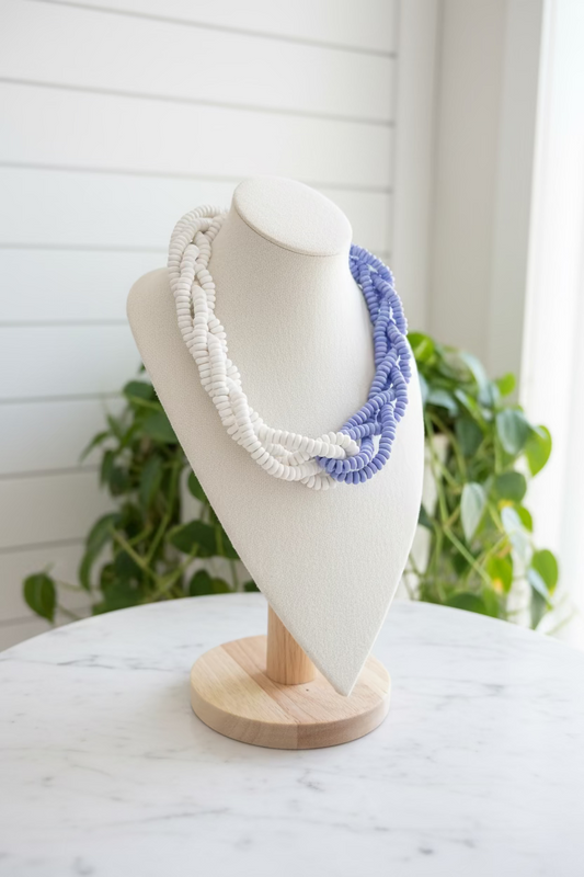 A multi-strand beaded necklace displayed on a cream-colored mannequin bust, featuring white beads on one side and lavender-blue beads on the other, set against a background of green houseplants and a wooden table.