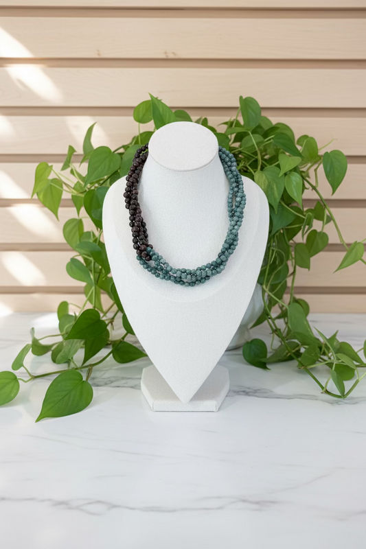 A multi-strand beaded necklace displayed on a white jewelry bust, featuring dark brown beads on one side and muted teal-green beads on the other, set on a wooden table with a plant in the background.