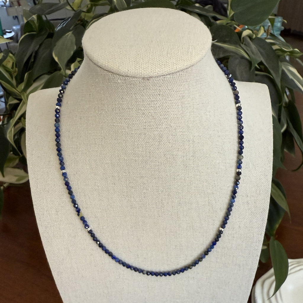 Blue beaded necklace on a white bust with greenery in the background