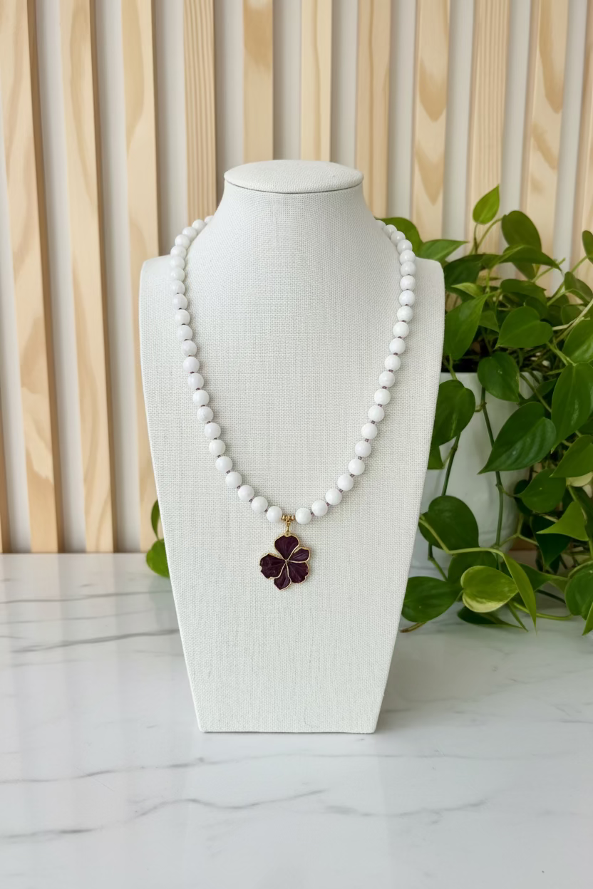 A white beaded necklace with a dark purple flower pendant, displayed on a cream-colored fabric mannequin bust on a wooden table with green plants in the background.