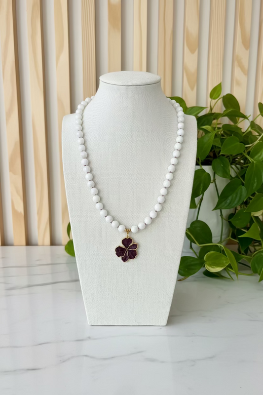 A white beaded necklace with a dark purple flower pendant, displayed on a cream-colored fabric mannequin bust on a wooden table with green plants in the background.