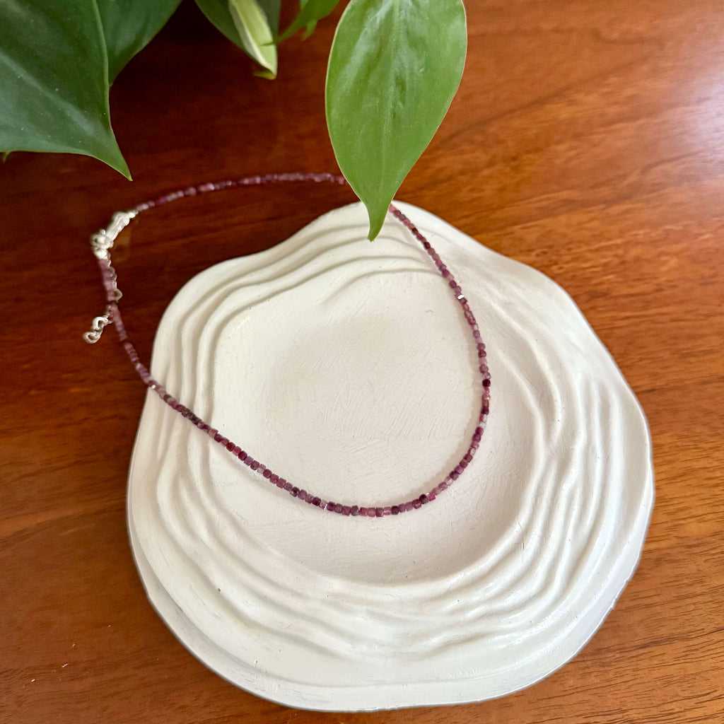Necklace with pink beads on a white dish on a wooden surface with a plant in the corner.
