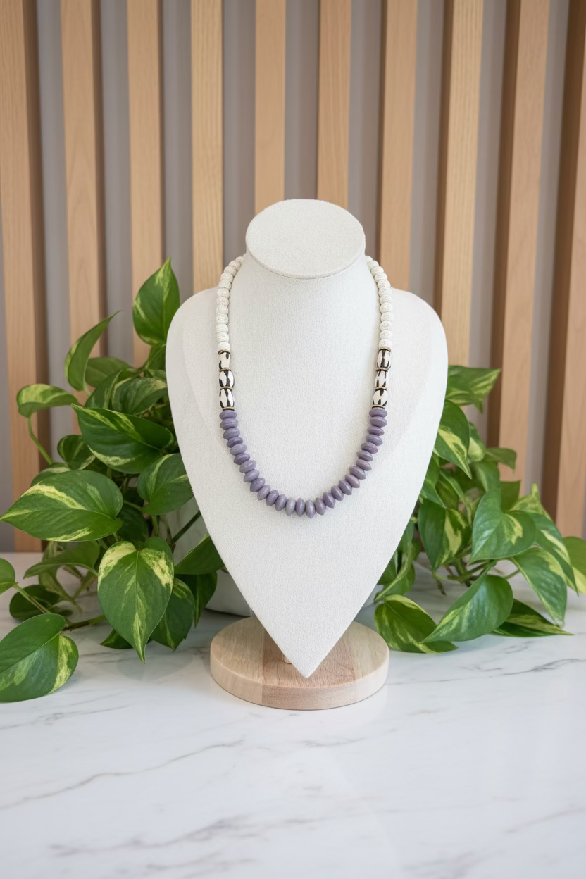 A beaded necklace displayed on a white fabric jewelry bust. The necklace features a top section of small white beads, a lower section of larger lavender-purple beads, and two metallic silver-toned spacer sections with black and white details.