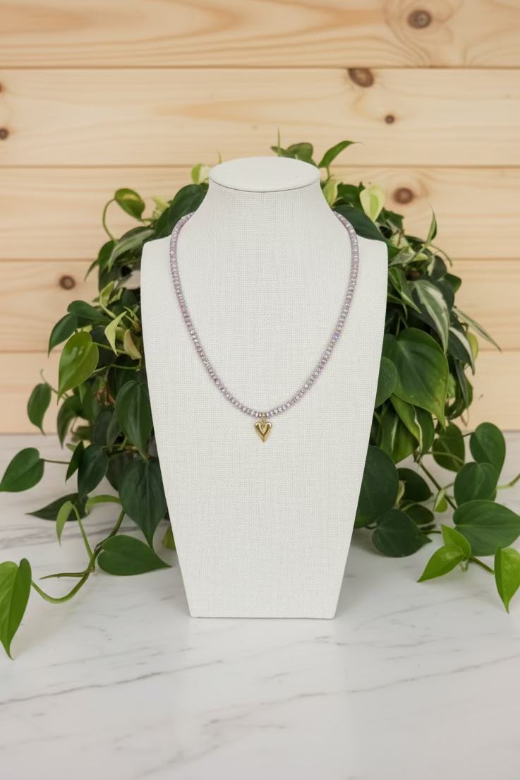A beaded necklace with iridescent lavender beads and a gold heart pendant, displayed on a cream fabric mannequin bust on a wooden table with green plants in the background.