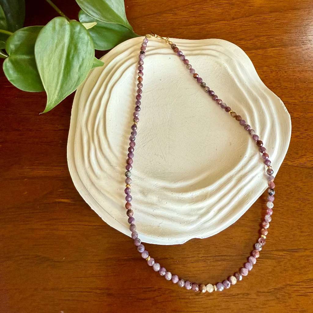 Necklace with purple beads on a white dish on a wooden surface with green leaves.
