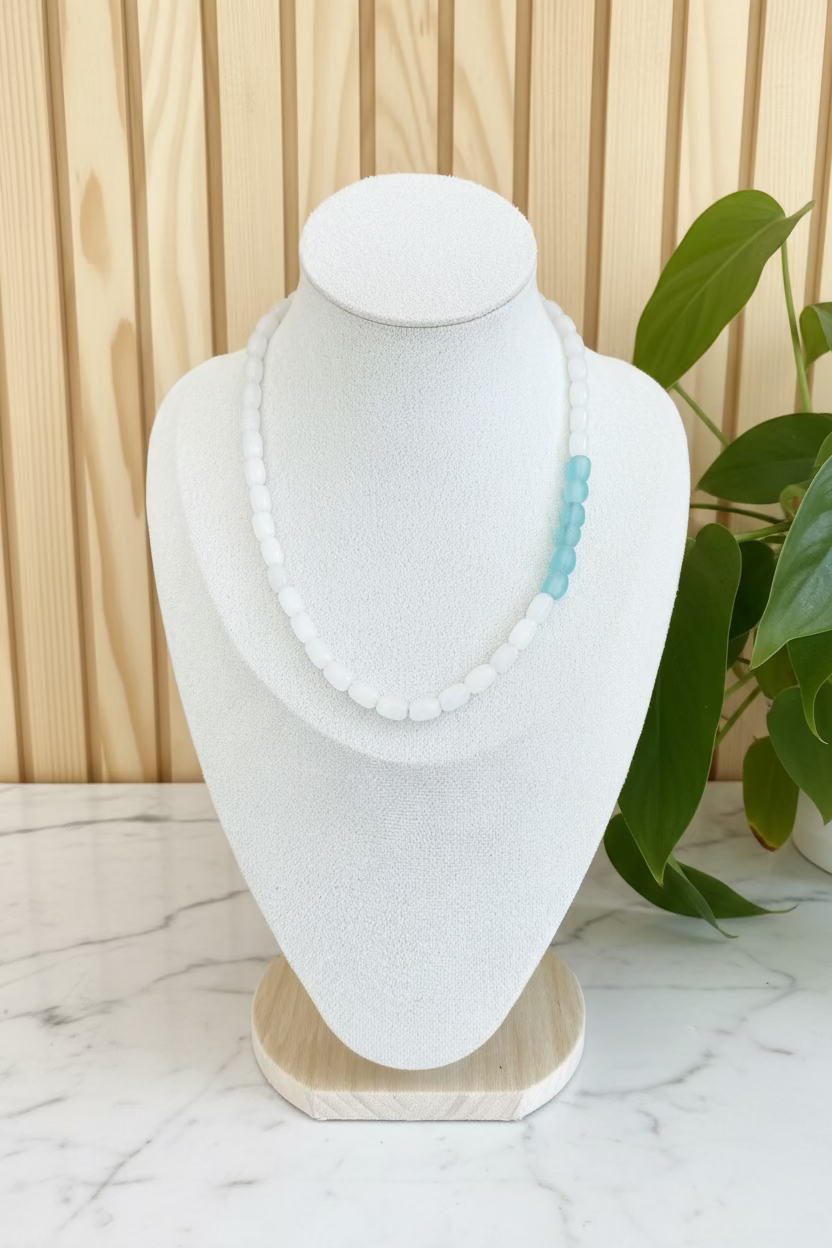A beaded necklace displayed on a textured white mannequin bust, featuring white round beads with a small cluster of light blue beads on one side, set against a background of indoor plants.