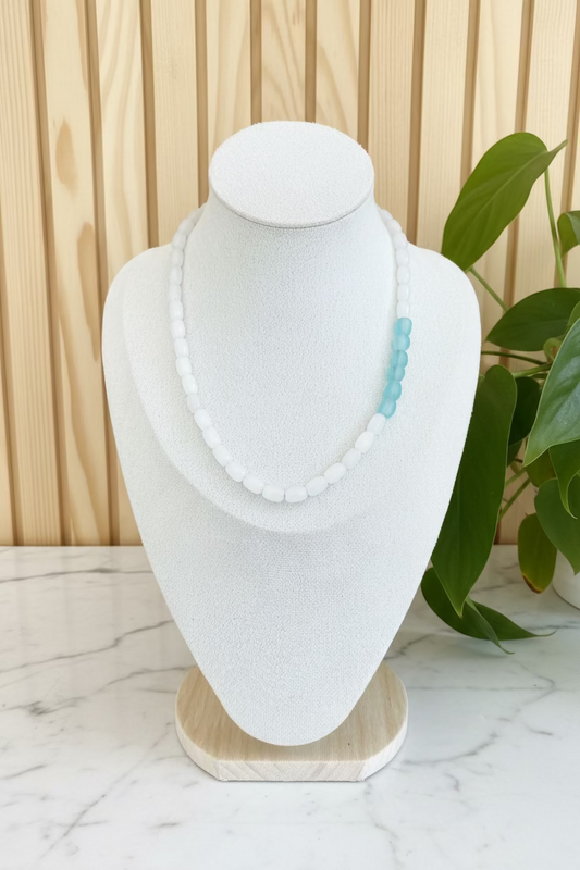 A beaded necklace displayed on a textured white mannequin bust, featuring white round beads with a small cluster of light blue beads on one side, set against a background of indoor plants.