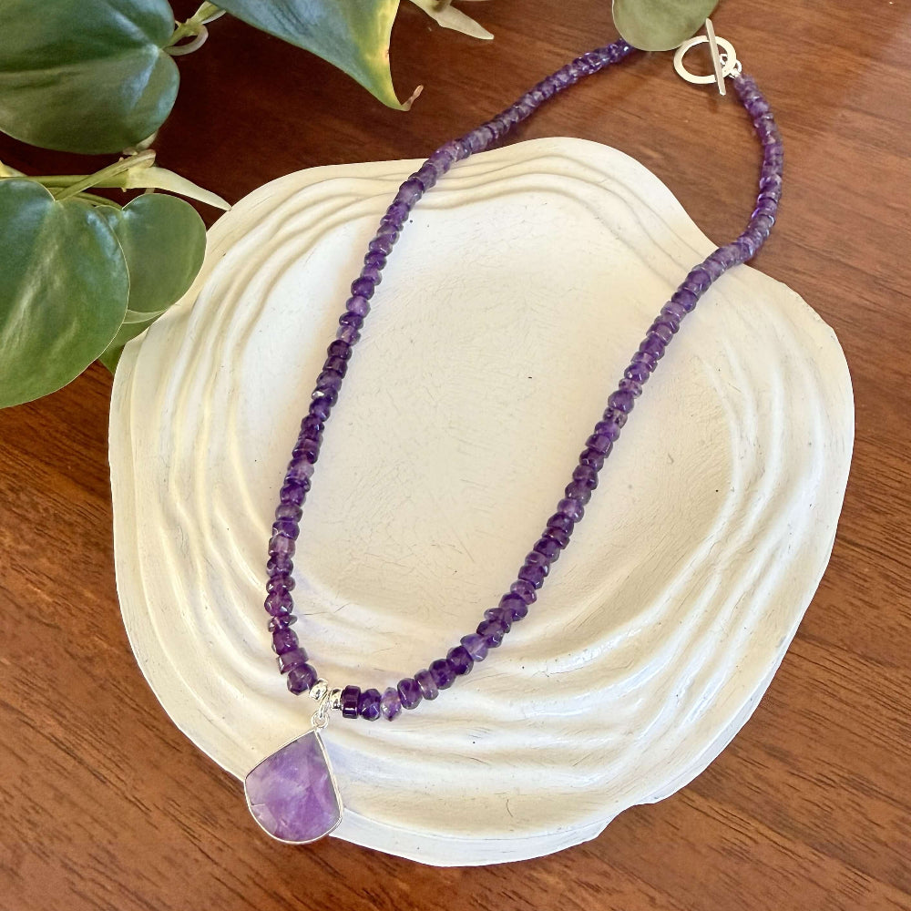 Purple amethyst beaded necklace with a purple pendant on a white shell with green leaves in the background