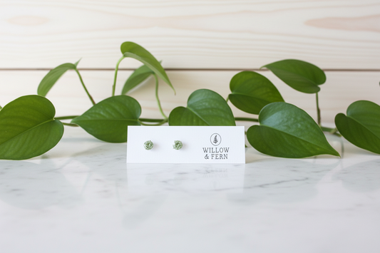 Two green stud aquamarine earrings on a Willow & Fern card sitting on a white marble counter with a plant in the background