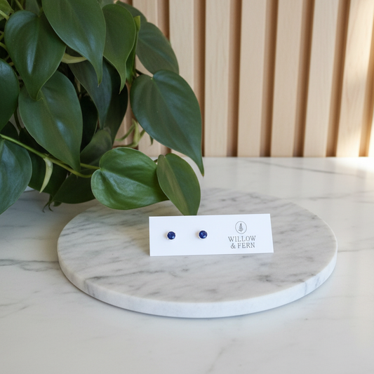 Two dark blue stud sapphire earrings on a Willow & Fern card sitting on a white marble counter with a plant in the background