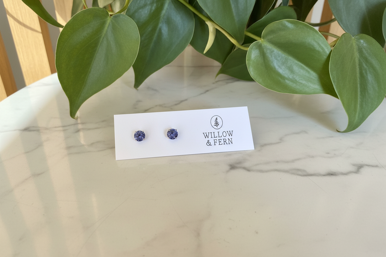 Two purple stud tanzanite earrings on a Willow & Fern card sitting on a white marble counter with a plant in the background
