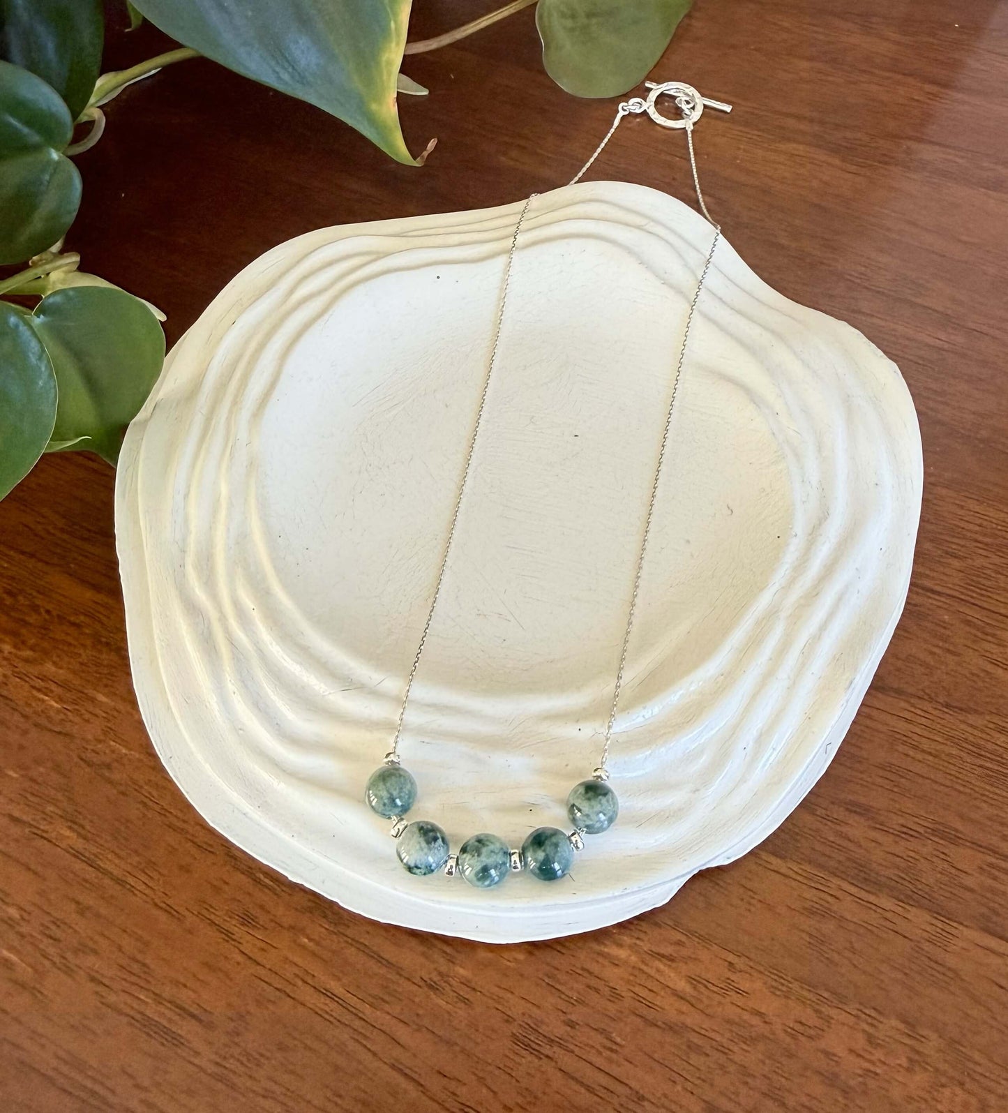 Jade beads on a silver necklace sitting on a white shell with green leaves in the background
