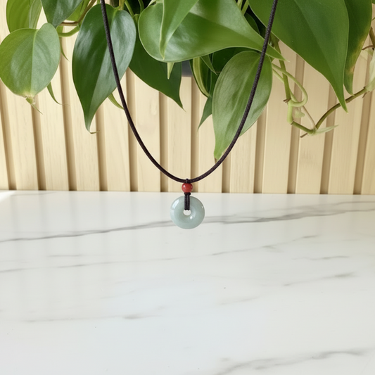 Necklace with a green jade pendant and a brown cotton cord on a white marble surface with a wooden background and a plant