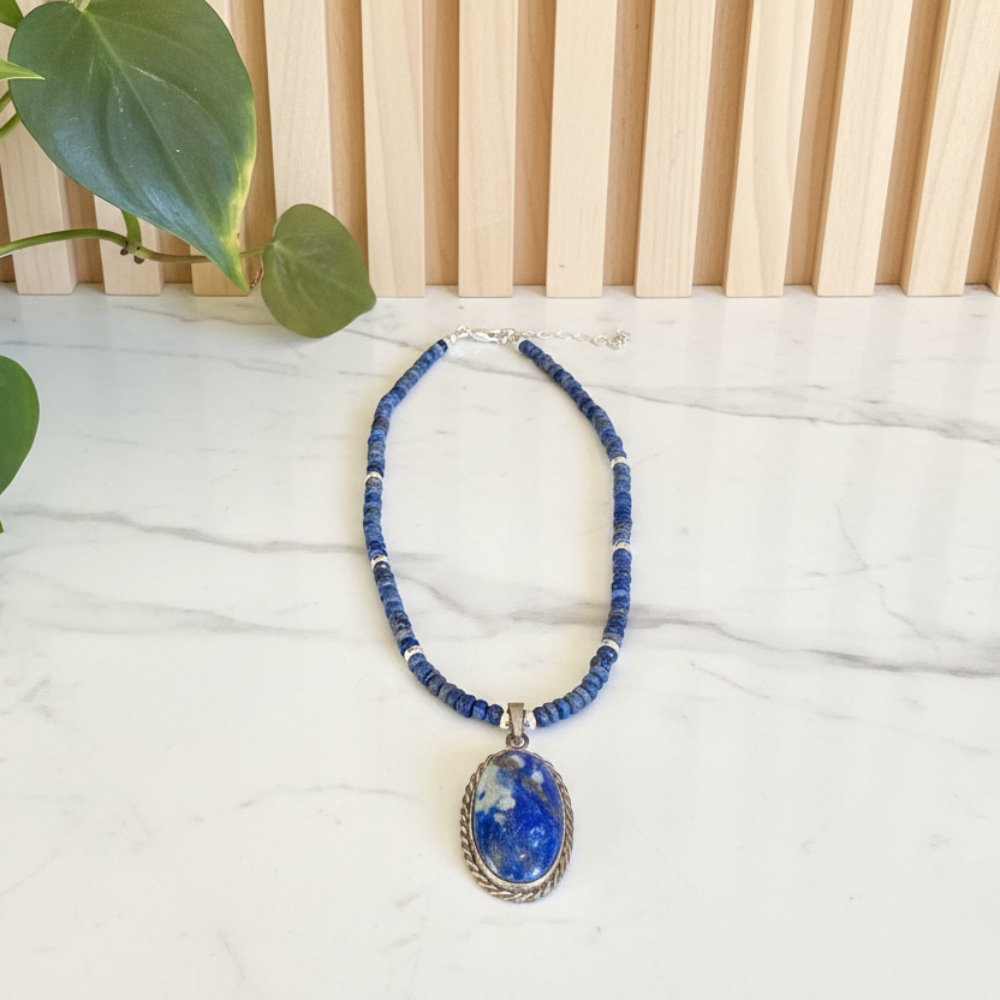 Blue lapis lazuli beaded necklace with a pendant on a white surface with a plant in the corner